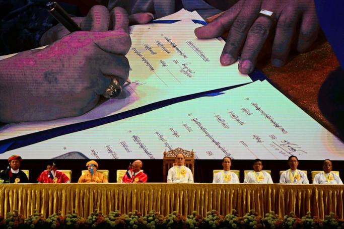 Myanmar president Thein Sein (C), leaders of ethnic armed groups (L) and government officials (R) sit on the stage as a leader of an ethnic armed group signs a document seen on a large screen (top) during a Nationwide Ceasefire Agreement (NCA) ceremony at the Myanmar International Convention Center (MICC) in Naypyitaw, Myanmar, 15 October 2015. Photo: Aung Htet/EPA
